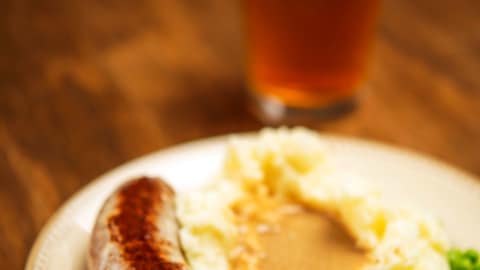 A plate of bangers sausage with mashed potatoes, peas and a beer.