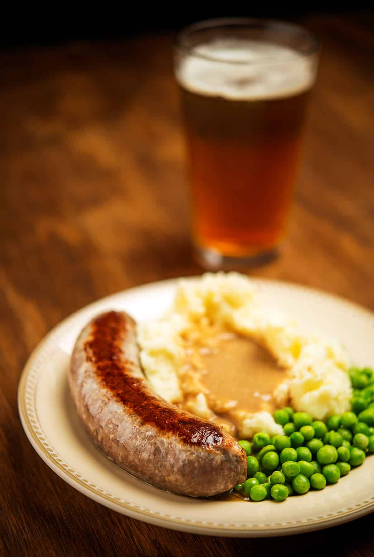 A plate of bangers sausage with mashed potatoes, peas and a beer. 