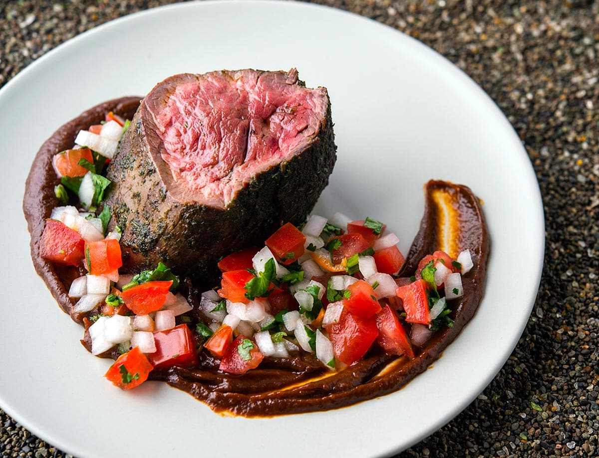 Seared elk tenderloin on a plate served with ancho sauce and pico de gallo. 