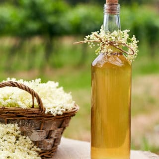 A bottle of elderflower liqueur with a basket of elderflowers next to it.