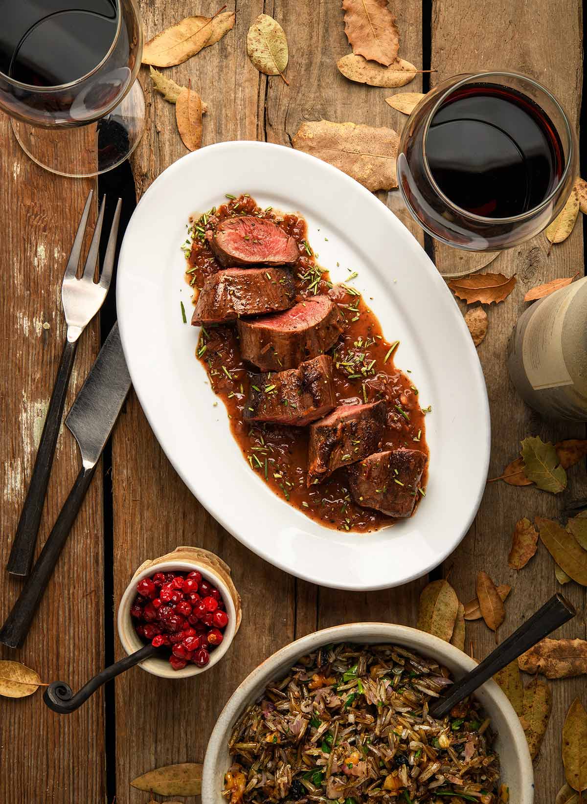 Pan seared venison tenderloin with a red wine sauce on a platter, alongside lingonberries and wild rice. 